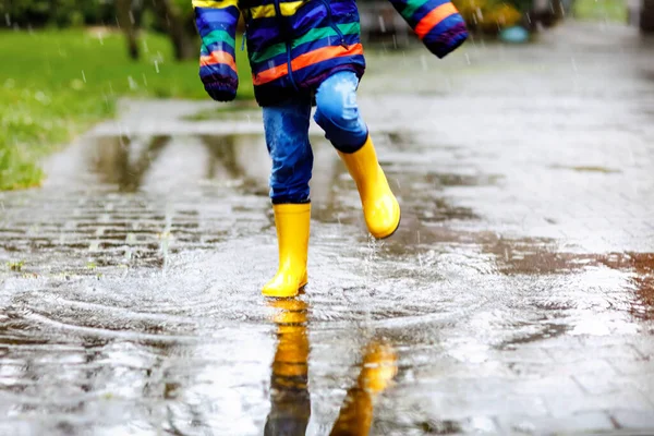 Primer plano del niño con botas de lluvia amarillas y caminando durante el aguanieve, la lluvia y la nieve en el día frío. Niño en ropa casual de moda colorida saltando en un charco. Divertirse al aire libre — Foto de Stock