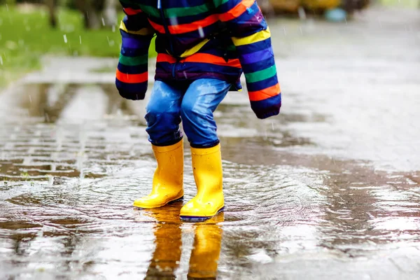 Close-up van het kind met gele regenlaarzen en wandelen tijdens ijzel, regen en sneeuw op koude dag. Kind in kleurrijke mode casual kleding springen in een plas. Veel plezier buiten — Stockfoto