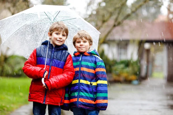 Two little kid boys on way to school walking during sleet, rain and snow with umbrella on cold day. Children, best friends and siblings in colorful fashion casual clothes — Stock Photo, Image