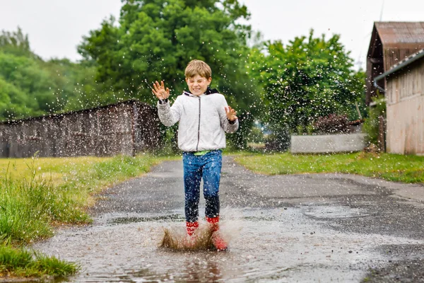 Niño feliz usando botas de lluvia rojas y caminando durante el aguanieve y la lluvia en el día nublado lluvioso. Niño con ropa casual de colores saltando en el charco. Divertirse al aire libre, actividad saludable para niños —  Fotos de Stock