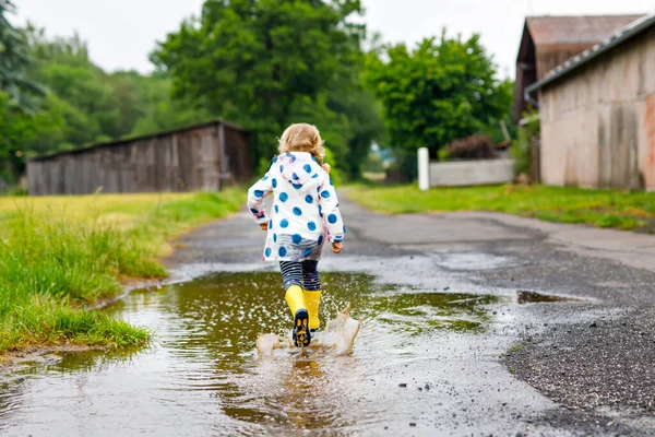 Маленька дівчинка в жовтих дощових черевиках бігає і ходить під час сну в дощовий хмарний день. Мила щаслива дитина в барвистому одязі, стрибає в калюжу, бризкається водою, активний відпочинок — стокове фото