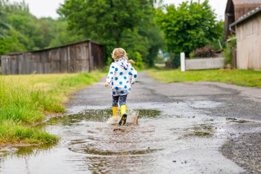 Küçük kız sarı çizme giyiyor, yağmurlu bir günde yağmurda yağmurda koşuyor ve yürüyor. Renkli giysiler içinde şirin mutlu bir çocuk su birikintisine atlıyor, su sıçrıyor, açık hava aktivitesi yapıyor.