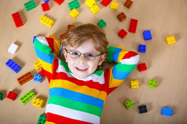 Piccolo bambino biondo con gli occhiali che gioca con un sacco di blocchi di plastica colorati al coperto. Ragazzo che indossa camicia colorata e divertirsi con la costruzione e la creazione — Foto Stock