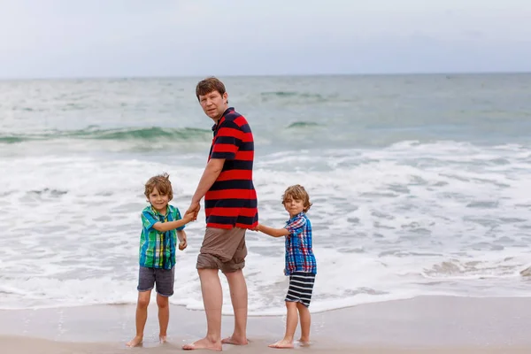 Due bambini felici ragazzi e papà in piedi sulla spiaggia dell'oceano e guardando all'orizzonte il giorno tempestoso. Famiglia, papà e figli carini fare vacanze, sognare e godersi l'estate — Foto Stock