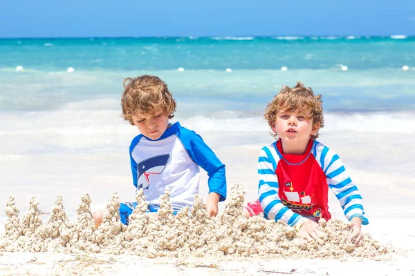 Dos niños divirtiéndose con la construcción de un castillo de arena en la playa tropical en la isla. Niños sanos jugando juntos en sus vacaciones. Gemelos, hermanos felices riendo y sonriendo — Foto de Stock