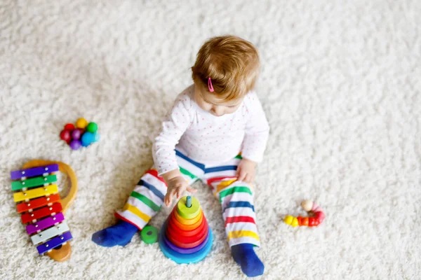 Адекватная симпатичная маленькая девочка, играющая с развивающими игрушками дома или в детском саду. Счастливый здоровый ребенок развлекается с разноцветной деревянной радужной игрушкой пирамидой. Ребенок учится различным навыкам — стоковое фото