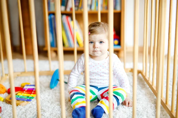 Beautiful little baby girl standing inside playpen. Cute adorable child playing with colorful toys. Home or nursery, safety for kids. Alone baby waiting for mom — Stock Photo, Image