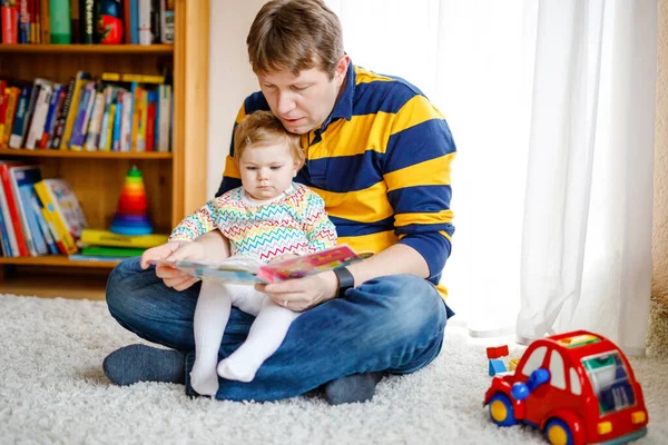 Genç baba, sevimli küçük kızıyla kitap okuyor. Gülümseyen güzel bir çocuk ve erkek evdeki oturma odasında birlikte oturuyorlar. Yeni yürümeye başlayan çocuk babaya kulak veriyor. — Stok fotoğraf