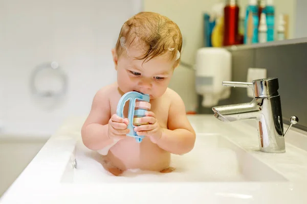 Schattige schattige baby nemen bad in wastafel en pak waterkraan. Klein gezond meisje met grote blauwe ogen hebben plezier en spelen met zeepschuim. — Stockfoto