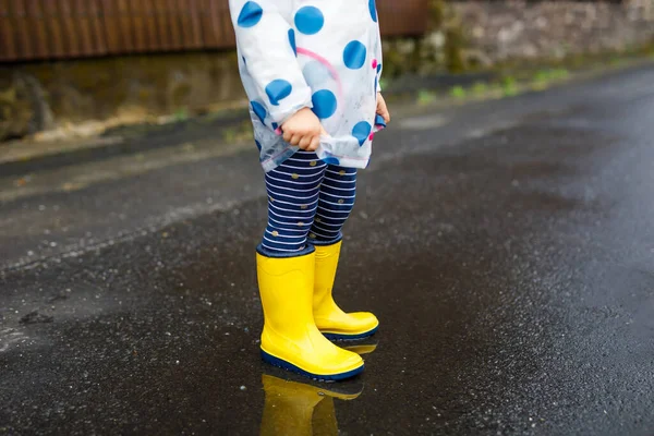 Primer plano de la niña pequeña con botas de lluvia amarillas y caminando durante el aguanieve en el día nublado lluvioso. Lindo niño en ropa colorida saltando en el charco, salpicaduras de agua, actividad al aire libre — Foto de Stock