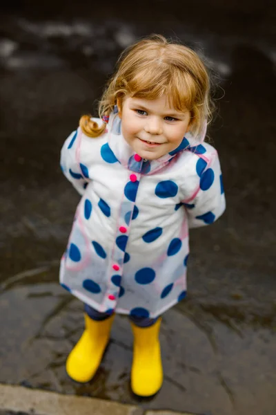 Menina pequena usando botas de chuva amarelas, correndo e andando durante o trenó em dia nublado chuvoso. Criança feliz bonito em roupas coloridas pulando em poça, salpicando com água, atividade ao ar livre — Fotografia de Stock