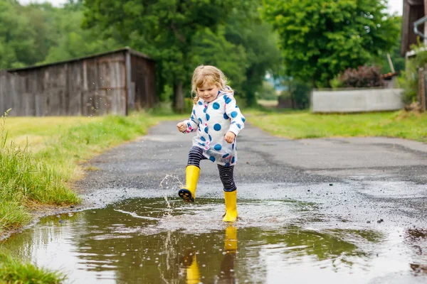 Kleines Mädchen in gelben Regenstiefeln, läuft und geht bei Schneeregen an einem regnerischen, bewölkten Tag. Nettes glückliches Kind in bunten Kleidern, das in eine Pfütze springt, mit Wasser planscht, Outdoor-Aktivität — Stockfoto