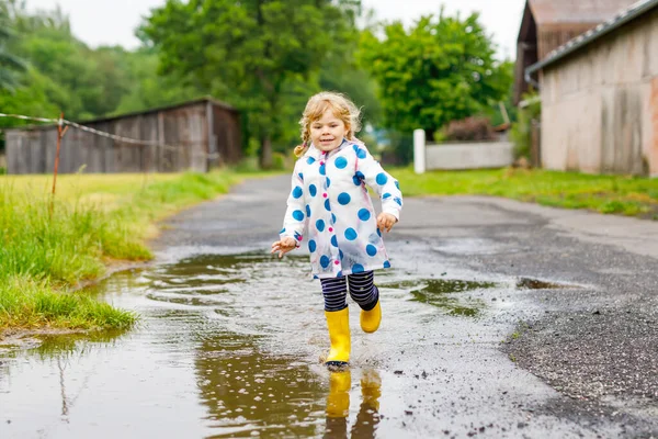 Niña pequeña con botas de lluvia amarillas, corriendo y caminando durante el aguanieve en el día nublado lluvioso. Lindo niño feliz en ropa colorida saltando en el charco, salpicaduras de agua, actividad al aire libre —  Fotos de Stock