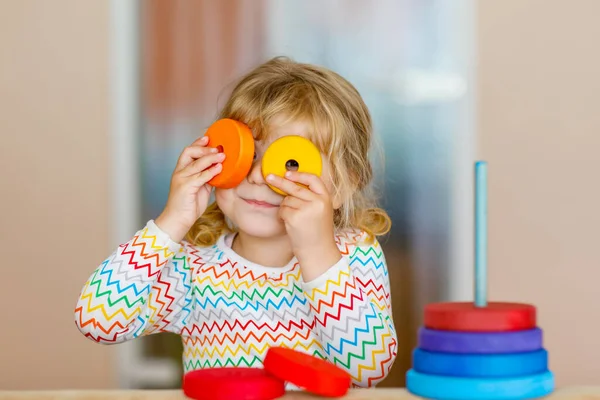 Симпатичная маленькая девочка, играющая одна с красочной деревянной радужной пирамидой и игрушками дома или в детской. Счастливый здоровый ребенок, развлекающийся в детском саду или дошкольном учреждении — стоковое фото