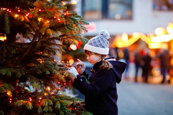 Little cute kid girl having fun on traditional Christmas market during strong snowfall. Happy child enjoying traditional family market in Germany. Schoolgirl standing by illuminated xmas tree. — Stock Photo, Image