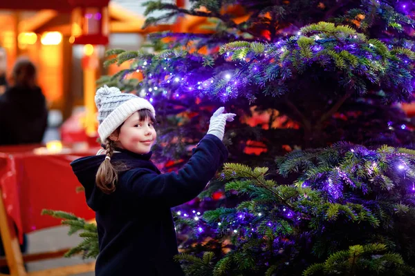 Malá roztomilá holčička baví na tradičním vánočním trhu během silného sněžení. Šťastné dítě těší tradiční rodinný trh v Německu. Školačka stojící ozářený vánoční strom. — Stock fotografie