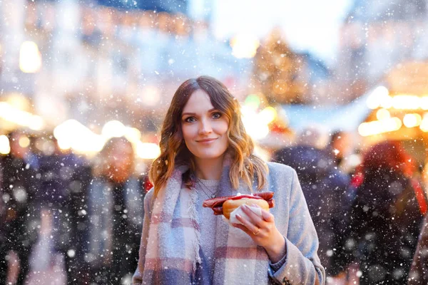 Güzel genç bir kadın, şiddetli kar yağışında geleneksel Noel pazarında eğleniyor. Sosis adı verilen geleneksel körili sosis yiyen mutlu kız. Aydınlatılmış Xmas ağacının yanında duran kadın.