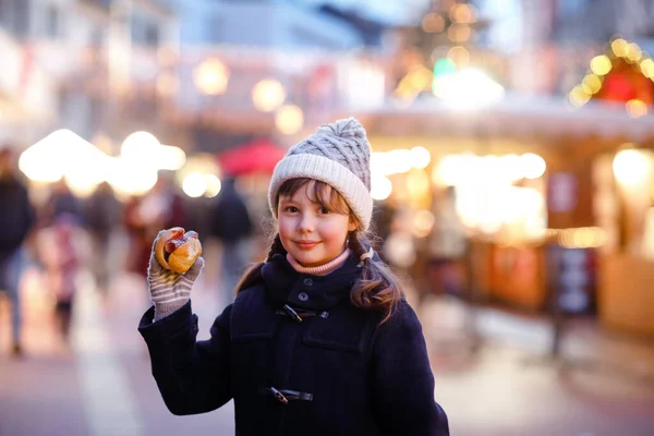 Cute little kid girl having fun on traditional Christmas market during strong snowfall. Happy child eating traditional curry sausage called wurst. schoolgirl standing by illuminated xmas tree. — Stock Photo, Image