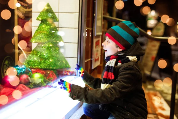 Cute little healthy school kid boy on Christmas market. Funny happy child in fashion winter clothes making window shopping decorated with gifts, xmas tree. Snow falling down, snowfall — Stock Photo, Image