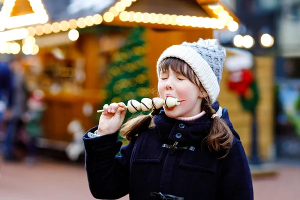 白いチョコレートを食べる小さなかわいい子供の女の子は、伝統的なドイツのクリスマスマーケットで串焼きにイチゴをカバーしました。雪の日にドイツの伝統的な家族市場で幸せな子供. — ストック写真