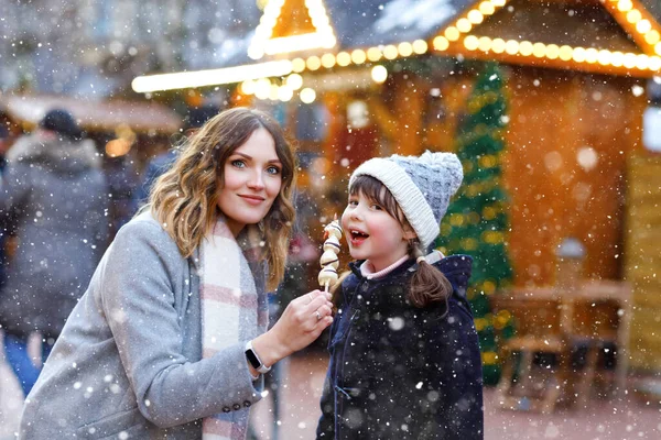 Madre e hija comiendo frutas cubiertas de chocolate blanco y fresa en brocheta en el tradicional mercado de Navidad alemán. Feliz chica y mujer en el mercado familiar tradicional en Alemania durante el día nevado —  Fotos de Stock