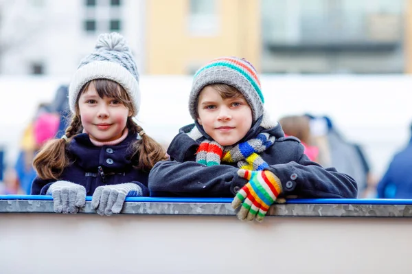 Zwei fröhliche kleine Mädchen und Jungen in bunten warmen Kleidern laufen auf einer Eisbahn des Weihnachtsmarktes oder Jahrmarktes. Gesunde Kinder haben Spaß auf Schlittschuhen. Viele Menschen mit aktiver Winterfreizeit. — Stockfoto