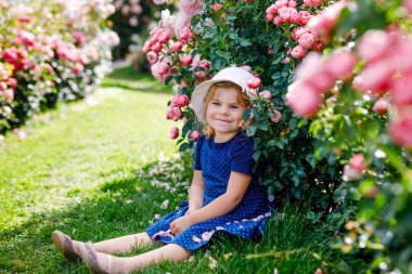 Çiçek açan gül bahçesinde küçük bir kızın portresi. Yaz güneşli bir günde parkta güllerle ve çiçeklerle eğlenen sevimli güzel bir çocuk. Mutlu gülümseyen bebek.