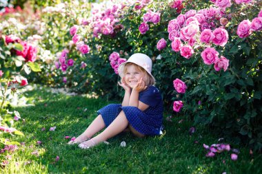 Çiçek açan gül bahçesinde küçük bir kızın portresi. Yaz güneşli bir günde parkta güllerle ve çiçeklerle eğlenen sevimli güzel bir çocuk. Mutlu gülümseyen bebek.