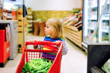 Süpermarkette alışveriş arabasını iten tatlı kız. Küçük çocuk meyve alıyor. Çocuk bakkal alışverişi. Tramvayı olan sevimli bebek marketten taze sebze seçiyor..