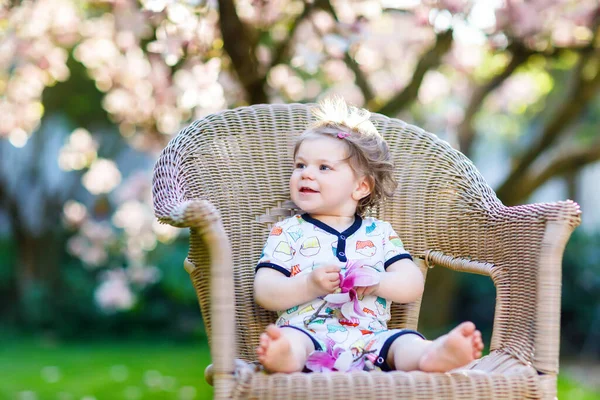 Söt liten flicka som sitter på en stor stol i trädgården. Vackra glada leende barn med blommande rosa magnolia träd på bakgrunden. Friska barn njuter av vårsäsongen. — Stockfoto