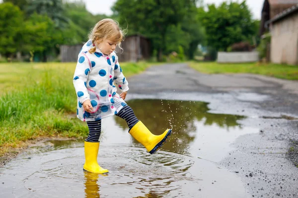 Liten småbarn flicka bär gula regnstövlar, springa och gå under slask på regnig molnig dag. Söt glad barn i färgglada kläder hoppar i pöl, stänk med vatten, utomhus aktivitet — Stockfoto