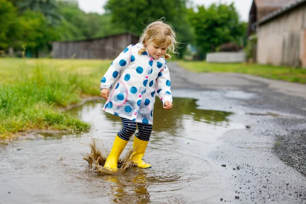 Petite fille en bas âge portant des bottes de pluie jaunes, courant et marchant pendant la neige fondante par temps de pluie nuageux. Enfant heureux mignon en vêtements colorés sautant dans la flaque d'eau, éclaboussures d'eau, activité de plein air — Photo