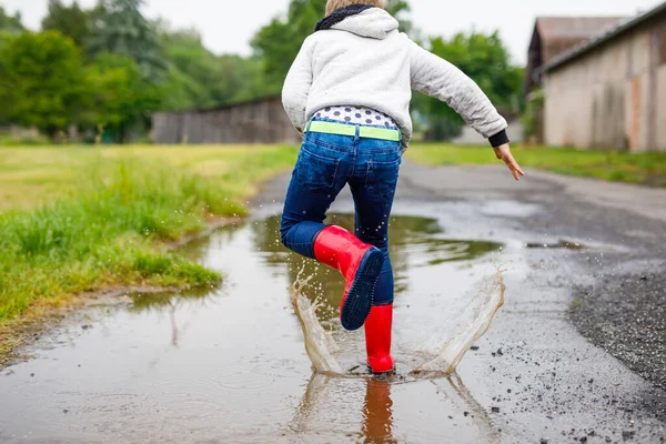 Menino feliz vestindo botas de chuva vermelha e andando durante a manga e chuva em dia chuvoso nublado. Criança em roupas casuais coloridas pulando na poça. Divirta-se ao ar livre, atividade de crianças saudáveis — Fotografia de Stock