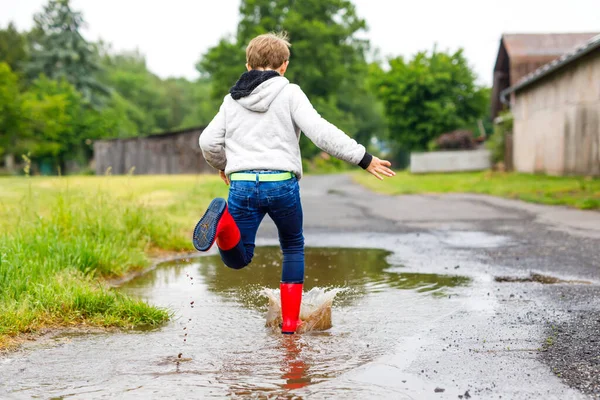 Happy kid boy wearing red rain boots and walking during sleet and rain on rainy cloudy day. Child in colorful casual clothes jumping into puddle. Having fun outdoors, healthy children activity — Stock Photo, Image