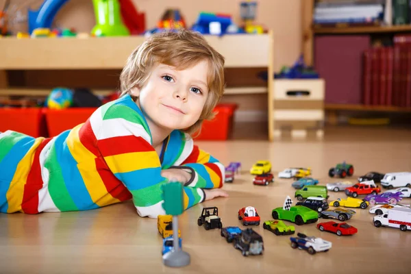 Härligt blond pojke leker med massor av leksaksbilar inomhus. Glad frisk pojke som har roligt under en pandemisk karantänsjukdom. Ensamstående barn hemma, stängd barnkammare. — Stockfoto