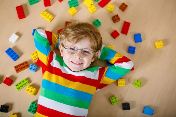 Little blond child with glasses playing with lots of colorful plastic blocks indoor. Kid boy wearing colorful shirt and having fun with building and creating — Stock Photo, Image