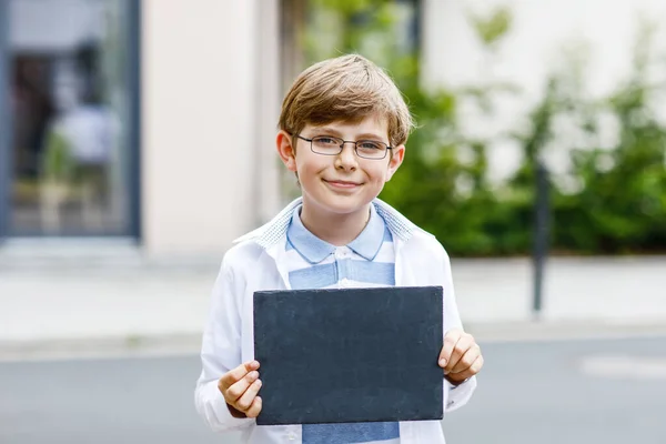 Glad liten pojke med glasögon och ryggsäck eller väska. Skolungen på väg till skolan. Friska förtjusande barn utomhus. Tomma krita skrivbord i händerna för copyspace och fri text. Tillbaka till skolan. — Stockfoto
