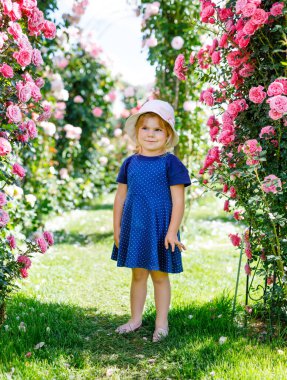 Çiçek açan gül bahçesinde küçük bir kızın portresi. Yaz güneşli bir günde parkta güllerle ve çiçeklerle eğlenen sevimli güzel bir çocuk. Mutlu gülümseyen bebek.