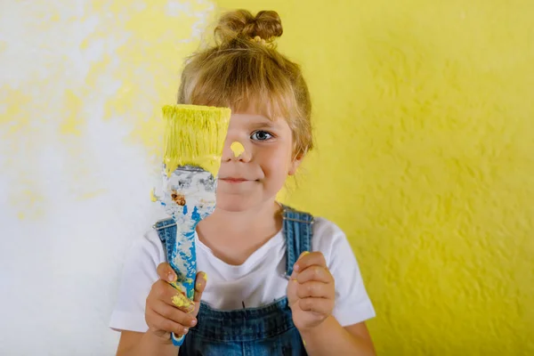 Симпатичная маленькая девочка, красит стену желтым цветом в новом доме. Семейный ремонт многоквартирного дома. Счастливая девочка рисует стены, выбирая цвет палитрой, развлекаясь кистью, в помещении — стоковое фото
