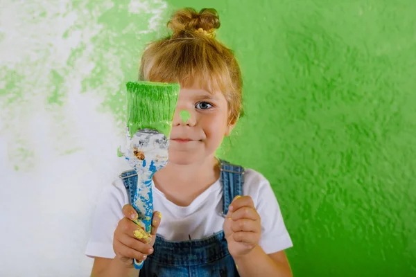 Söt liten småbarn flicka måla väggen med gul färg i nytt hus. Familjen reparerar lägenheten hemma. Glad barn flicka målar väggen, välja färg med palett, ha kul med borste, inomhus — Stockfoto