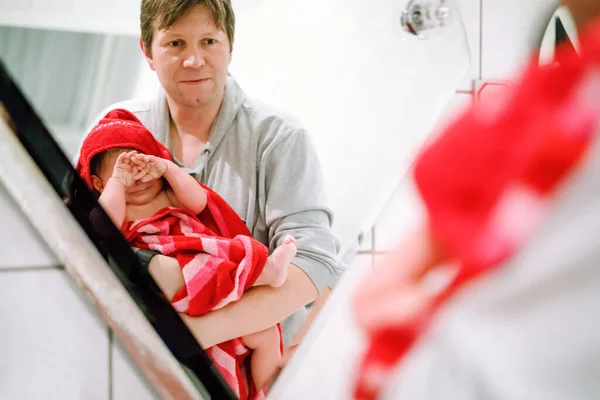 Father holding cute little toddler girl wrapped in towel after taking bath. Happy healthy baby child with wet hairs looking in mirror in bathroom and laughing. Family relationship of daughter and dad — Stock Photo, Image