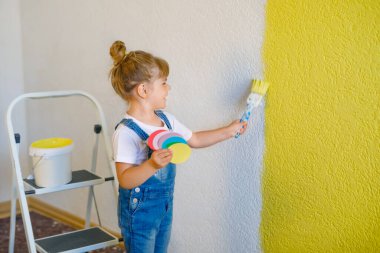 Küçük şirin kız yeni evdeki duvarları sarı renkle boyuyor. Aile onarım dairesi. Mutlu çocuk duvarı boyuyor, paletle renk seçiyor, fırçayla eğleniyor, kapalı alanda.