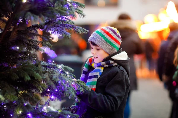 Pequeno garoto bonito menino se divertindo no mercado de Natal tradicional durante forte queda de neve. Criança feliz desfrutando do mercado familiar tradicional na Alemanha. Estudante em pé junto à árvore de xmas iluminada. — Fotografia de Stock