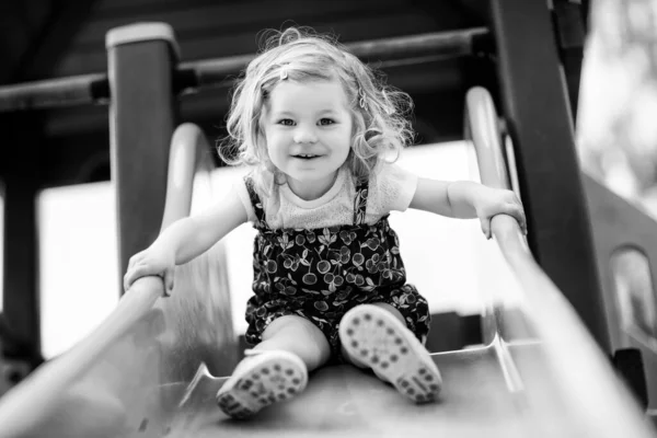 Petite fille mignonne jouant sur la glissière sur l'aire de jeux extérieure. Beau bébé en short coloré qui s'amuse par une chaude journée d'été ensoleillée. Enfant glissant vers le bas — Photo
