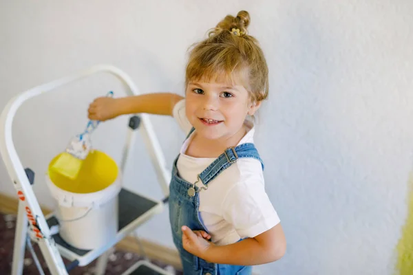 Menina pequena adorável da criança pintando a parede com cor na casa nova. Casa de apartamento de reparação familiar. Criança bebê feliz pinta a parede, escolhendo a cor com paleta, se divertindo com escova, dentro de casa — Fotografia de Stock