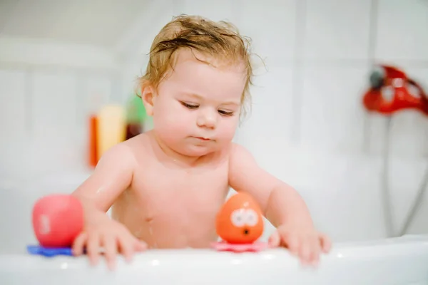 Cute adorable baby girl taking foamy bath in bathtub. Toddler playing with bath rubber toys. Beautiful child having fun with colorful gum toys and foam bubbles — Stock Photo, Image