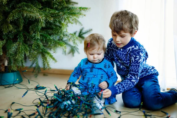 Χαριτωμένο κοριτσάκι και μικρό αγόρι κατεβάζουν χριστουγεννιάτικα στολίδια από το χριστουγεννιάτικο δέντρο. παιδιά, αδέλφια, αδελφός και αδελφή κρατώντας ελαφρά γιρλάντα. Οικογένεια μετά τον εορτασμό αφαιρέστε και απορρίψτε δέντρο — Φωτογραφία Αρχείου
