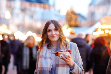 Güzel genç bir kadın, şiddetli kar yağışında geleneksel Noel pazarında eğleniyor. Sosis adı verilen geleneksel körili sosis yiyen mutlu kız. Aydınlatılmış Xmas ağacının yanında duran kadın.