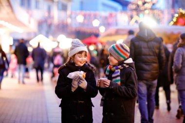 Cute little kids girl and boy having fun on traditional Christmas market during strong snowfall. Happy children eating traditional curry sausage called wurst and drinking hot chocolate. Twins friends clipart