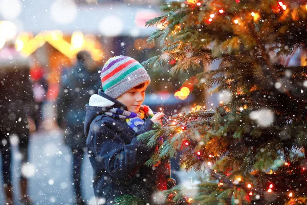 Little cute kid boy having fun on traditional Christmas market during strong snowfall. Happy child enjoying traditional family market in Germany. Schoolboy standing by illuminated xmas tree. — Stock Photo, Image